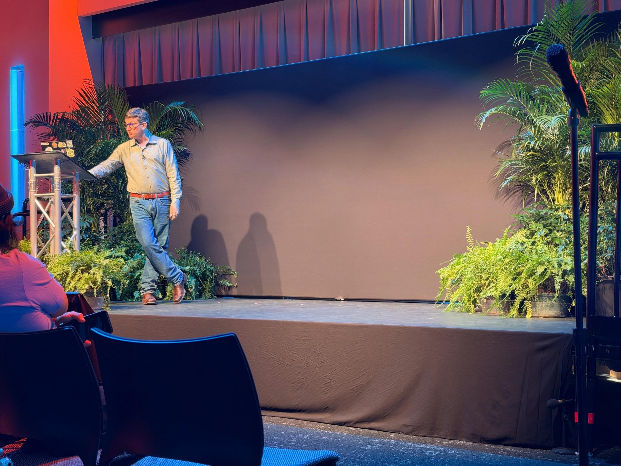 Corey Quinn presenting his keynote on a stage framed by plants.
